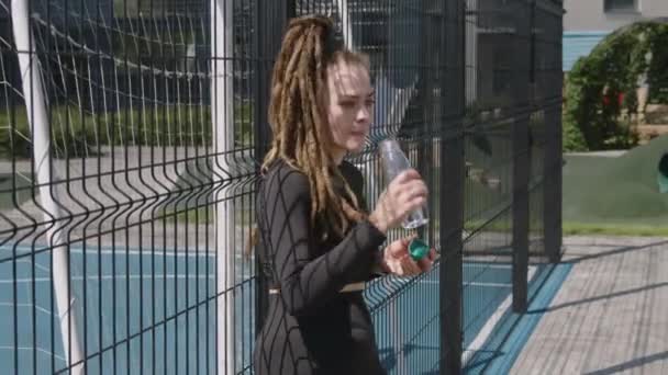Side felvétel kaukázusi vonzó női sportoló raszta ivóvíz edzés után, miközben támaszkodik a kerítés rekreációs terület napsütéses napon - Felvétel, videó