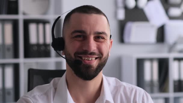 Muotokuva hymyilevä valkoihoinen mies hotline operaattori kuulokkeet työskentelevät toimistossa, katsot kameraa. Televiestintä, telemarkkinointi, neuvontapuhelin, online-tuki. - Materiaali, video