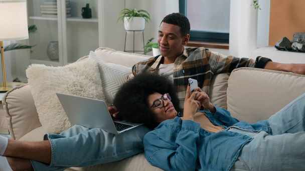 Afro-Américain couple homme avec ordinateur portable assis sur canapé maison femme bavarder smartphone navigation téléphone mobile pose sur les genoux mâle fille gars gadget addiction internet sourire famille - Photo, image