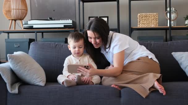 Кавказька мати та її маленький син використовують мобільний телефон для перегляду відео в Інтернеті, сидячи разом на дивані у вітальні. Онлайн мультфільми або освітні відео. - Кадри, відео