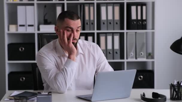 Yorgun ve uykulu Kafkasyalı iş adamı laptopla çalışıyor, ofisinde oturuyor. Duygusal stres, iş sorunları, can sıkıntısı veya tükenme kavramı. - Video, Çekim