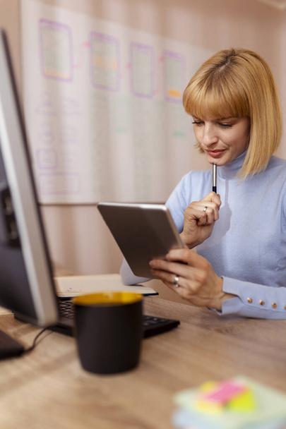 Woman sitting at her desk in home office, taking an online course using tablet computer, writing in a planner - Zdjęcie, obraz