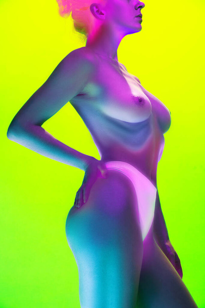 Young woman in innerwear with naked body, breast in blue-magenta neon light against gradient green background. Concept of art and femininity, female health, body care, self-expression and self-love. - Photo, Image
