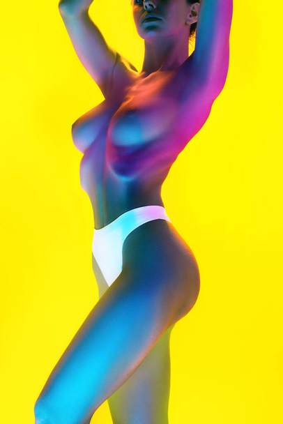 Young elegant, naked woman raising hands in magenta-blue neon light against gradient yellow background. Model with perfect curves. Concept of femininity, female health, self-expression and self-love. - Photo, Image