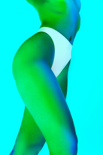 Oříznutý portrét ženského modelu s štíhlým, nahým tělem v zeleném neonovém světle na svahu modrého pozadí. Koncept umění a ženskosti, zdraví žen, péče o tělo, sebevyjádření a sebelásky. - Fotografie, Obrázek