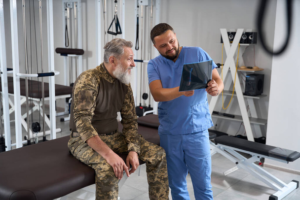 Fyzioterapeut s pacientem ve vojenském oděvu vyšetřuje rentgeny, oba muži jsou vousatí - Fotografie, Obrázek