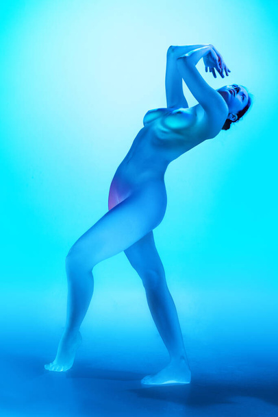 Jonge naakte vrouw dansend in neon levendig licht tegen blauwe gradiënt achtergrond. Model met perfecte body curves. Concept van kunst en vrouwelijkheid, vrouwelijke gezondheid, lichaamsverzorging, zelfexpressie en eigenliefde - Foto, afbeelding