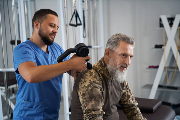 Fyzioterapeut provádí hardwarovou masáž na zádech vojáka během rehabilitace, pacienta v maskovacím oděvu - Fotografie, Obrázek