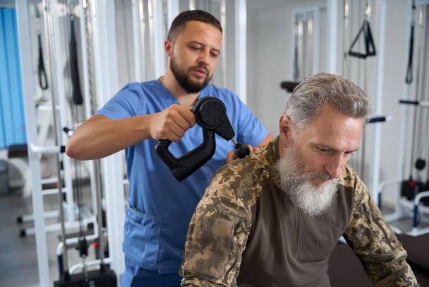 Врач делает аппаратный массаж на спине военного во время реабилитации, пациент в камуфляжной одежде - Фото, изображение