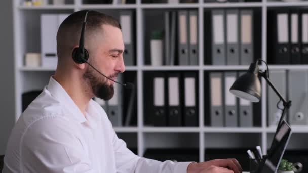 Kafkas erkek temas merkezi çalışanı kulaklık takıyor ve dizüstü bilgisayar kullanıyor, müşteriyle konuşuyor, ofiste çalışıyor. Çevrimiçi yardım veya teknik destek sağlanması. - Video, Çekim