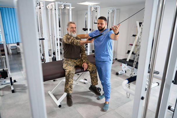 Trainer helping military patient work on exercise machine, man doing muscle stretching exercises - Фото, изображение