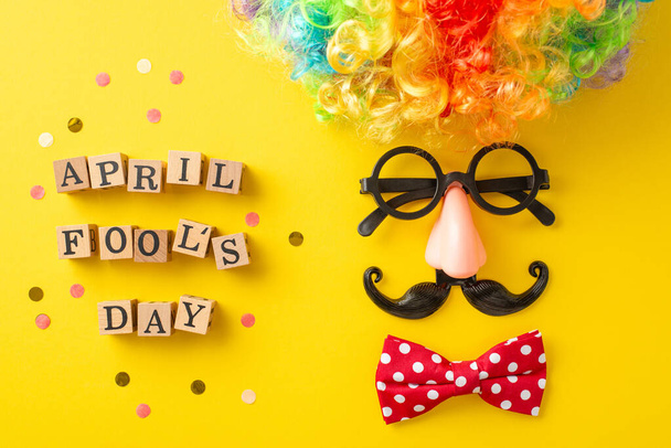Top-down foto van "April Fool 's Day" in blokletters, met een clown' s accessoires, waaronder een pruik, strikje, bril met valse neus, en snor, gerangschikt als gezicht op gele achtergrond met confetti - Foto, afbeelding