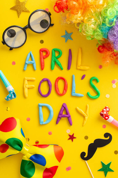 Verticale foto van bovenaf, met "April Fool 's Day" in kleiletters, stripbril, pruik van de nar, kleurrijke strikje, valse snor, blaashoorns en glitter op een levendige gele achtergrond - Foto, afbeelding