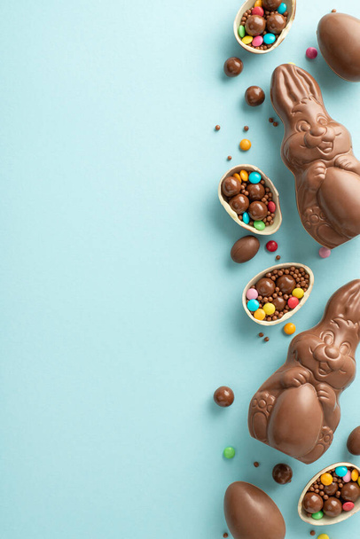 Adorable Easter ensemble idea. Vertical top view of cracked chocolate eggs, filled with colorful candies, a chocolate bunnies, sprinkles on a pastel blue background, placeholder for text or advert - Photo, Image