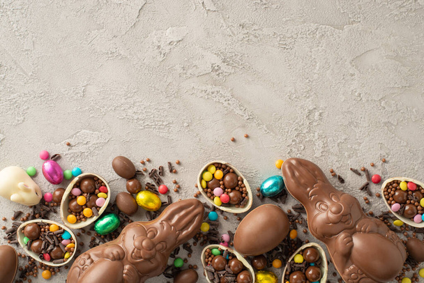 Modern Paskalya şekeri sunumu. Renkli şekerlerle dolu kırık çikolata yumurtalarının tepeden tırnağa görüntüsü, çikolatalı tavşanlar, ve dokulu gri arkaplan üzerine serpiştirilmiş serpiştirilmiş, mesaj için geniş bir alan. - Fotoğraf, Görsel
