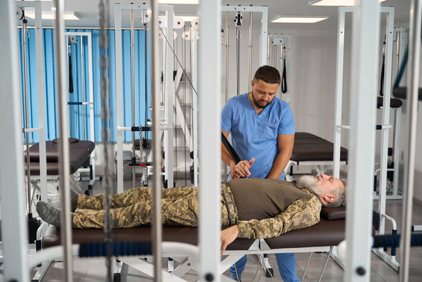 Военный пациент делает упражнения, чтобы растянуть мышцы рук на специальном тренажере, ему помогает реабилитатор - Фото, изображение