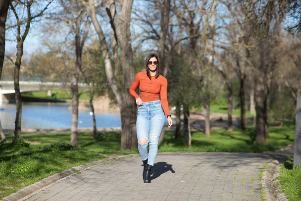 Nuori ja kaunis brunette nainen pukeutunut rento vaatteita ja aurinkolasit, kävelee polkua lehtipuiden välillä syksyllä, taustalla järvi - Valokuva, kuva