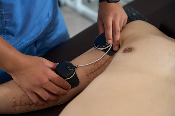 Rehabilitasyon uzmanı elektrotları erkeğin koluna bağlar, hasta masaj masasında yatar. - Fotoğraf, Görsel