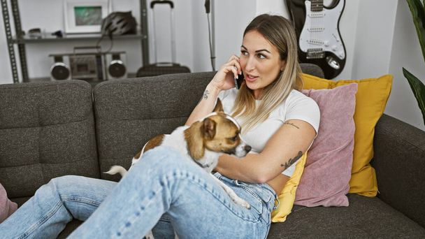 Μια νεαρή γυναίκα μιλάει στο τηλέφωνο ενώ αγκαλιάζει το σκύλο της σε έναν καναπέ σε ένα άνετο σαλόνι. - Φωτογραφία, εικόνα