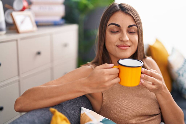 Giovane bella donna ispanica che beve caffè seduta sul divano a casa - Foto, immagini