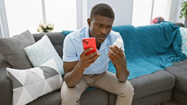 Hombre afroamericano usando teléfono inteligente y sosteniendo la botella de medicina mientras está sentado en el sofá en el interior - Foto, Imagen