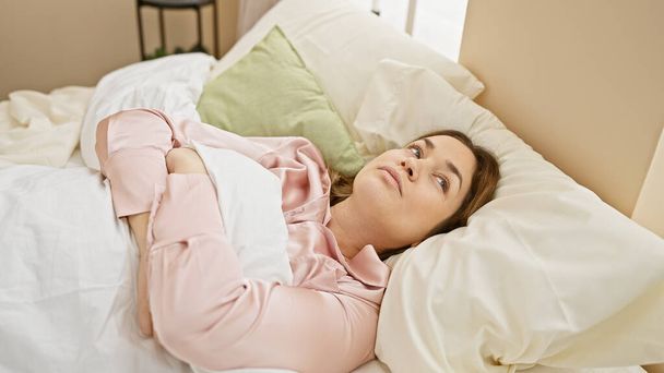 Пухнаста молода жінка з брюнеткою волоссям і блакитними очима в рожевій блузці, лежачи в ліжку, в приміщенні. - Фото, зображення