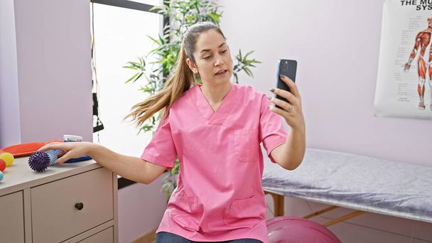 Жінка в рожевих скрабах тримає телефон для відеодзвінка в терапевтичній кімнаті з реабілітаційним обладнанням. - Фото, зображення