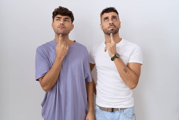 Homosexuelles homosexuelles Paar steht vor weißem Hintergrund und denkt konzentriert über Zweifel nach, mit dem Finger am Kinn und schaut fragend nach oben  - Foto, Bild