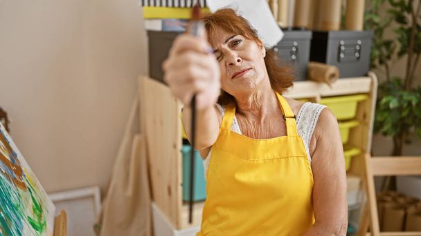 A mature woman in a yellow apron focuses intently while painting on a canvas in an art studio. - Photo, Image