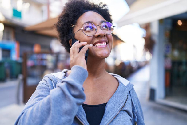 Mujer afroamericana sonriendo confiada hablando en el teléfono inteligente en la terraza de la cafetería - Foto, Imagen