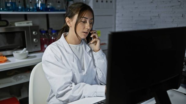 Jonge volwassen Latijns-Amerikaanse vrouw draagt laboratoriumjas binnen, met behulp van computer en telefoon in het laboratorium - Foto, afbeelding