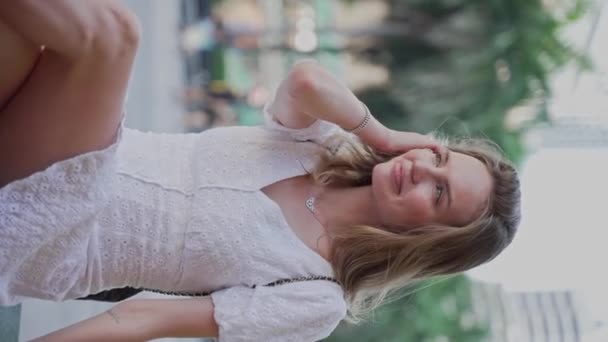 Adorable joven mujer caucásica en vestido blanco de verano sentado en el parque de la ciudad, mirando a la cámara y sonriendo - Imágenes, Vídeo