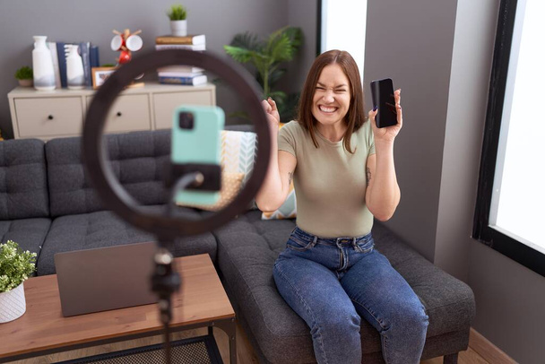 Beautiful brunette woman recording vlog tutorial with smartphone at home screaming proud, celebrating victory and success very excited with raised arm  - Photo, Image