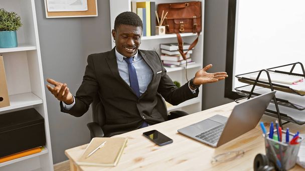 近代的なオフィスでノートパソコンとスマートフォンで机に座っている間,自信を持ったアフリカのビジネスマンがジェスチャー. - 写真・画像