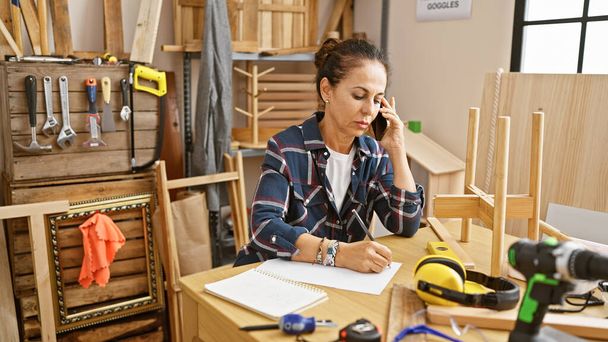 Una mujer hispana de mediana edad en un taller hablando por teléfono mientras está rodeada de herramientas de carpintería y muebles. - Foto, Imagen