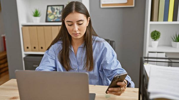 Focused hispanic woman using laptop and smartphone in modern office setting - Photo, Image