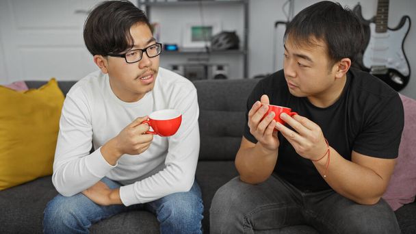 Dva asijští muži mají rozhovor v moderním obývacím pokoji, zatímco drží červené šálky kávy - Fotografie, Obrázek