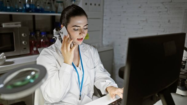 Μια γυναίκα επιστήμονας πολλαπλών καθηκόντων σε ένα εργαστήριο, ενώ μιλάμε σε ένα τηλέφωνο και χρησιμοποιώντας έναν υπολογιστή. - Φωτογραφία, εικόνα