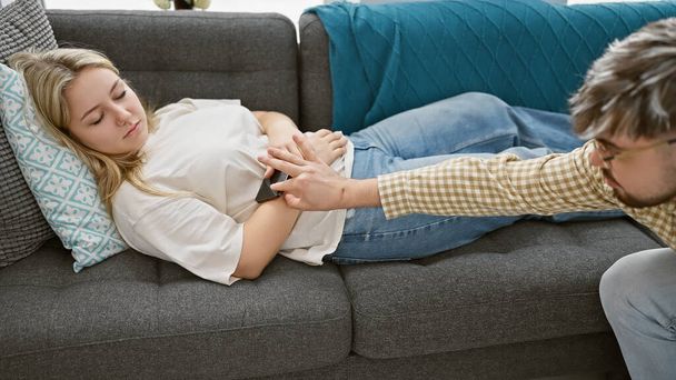 En un ambiente interior, un hombre recupera sigilosamente un teléfono de la mano de una mujer dormida en un sofá. - Foto, imagen
