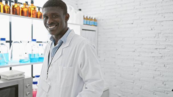 Lächelnder afrikanischer Mann in weißem Laborkittel steht in einem Labor mit Bechern und Fläschchen und vermittelt Professionalität und Gesundheit. - Foto, Bild