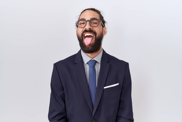 Homem hispânico com barba vestindo terno e gravata colando a língua feliz com expressão engraçada. conceito de emoção.  - Foto, Imagem