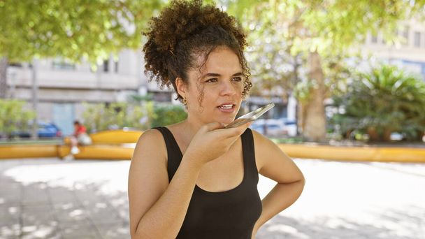 Молода іспаномовна жінка з кучерявим волоссям записує голосове повідомлення на своєму смартфоні на відкритому повітрі в міському парку. - Фото, зображення