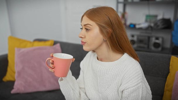 Zrelaksowana młoda kobieta pije filiżankę kawy na wygodnej sofie w nowoczesnym salonie. - Zdjęcie, obraz