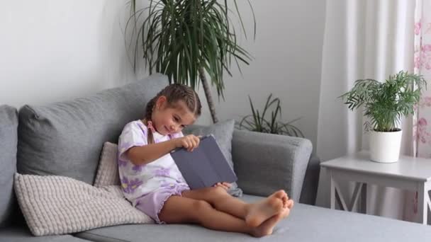 Suloinen pieni tyttö istuu kotona sohvalla ja pelata pelejä tabletilla. Kotituulella. Laadukas kuva - Materiaali, video