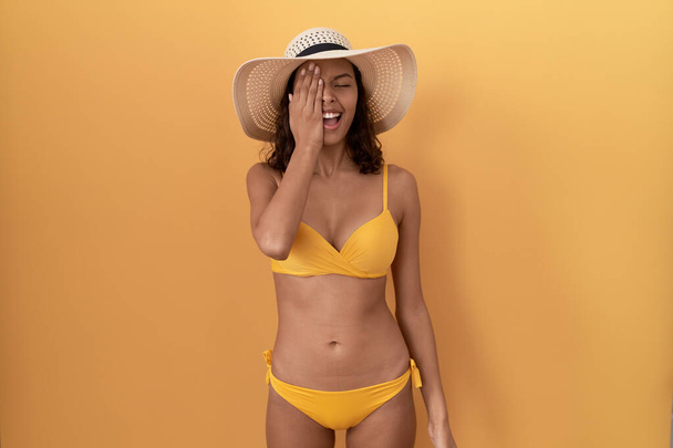 Young hispanic woman wearing bikini and summer hat covering one eye with hand, confident smile on face and surprise emotion.  - Foto, Bild