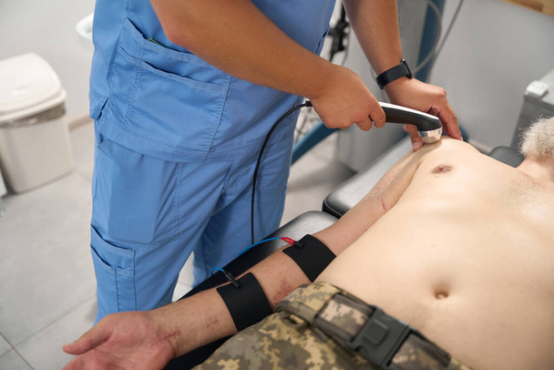 Militaire man met littekens op zijn lichaam tijdens een hardware therapie sessie, de arts maakt gebruik van een modern apparaat - Foto, afbeelding