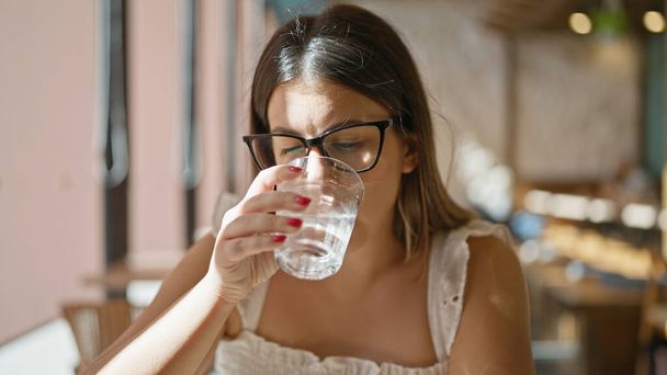 Mooie jonge Spaanse vrouw draagt een bril en drinkt een glas water in een cafetaria - Foto, afbeelding