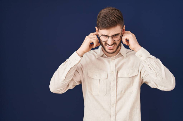 Іспаномовний чоловік з бородою стоїть над синім тлом, покриваючи вуха пальцями з роздратованим виразом для шуму гучної музики. глуха концепція.  - Фото, зображення