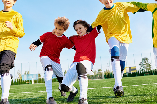 Μια ομάδα μικρών παιδιών με πολύχρωμες φανέλες τρέχουν, κλωτσάνε, και πασάρουν μια μπάλα ποδοσφαίρου σε ένα χορταστικό πεδίο κάτω από τον λαμπερό ήλιο. - Φωτογραφία, εικόνα