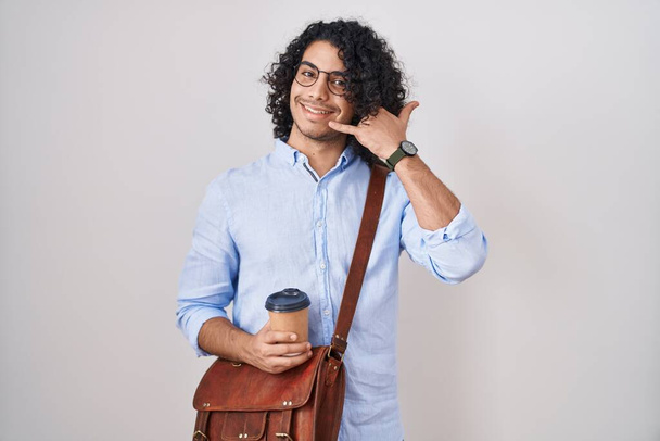 Homem hispânico com cabelo encaracolado bebendo uma xícara de café sorrindo fazendo gesto de telefone com a mão e os dedos, como falar ao telefone. conceitos comunicantes.  - Foto, Imagem